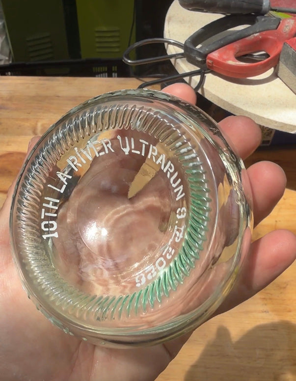 A hand holding a clear glass jar lid with embossed white text reading '10TH LA RIVER ULTRARUN 2026' and green spiral detailing around the rim, shown against a workshop background with tools and a wooden surface.