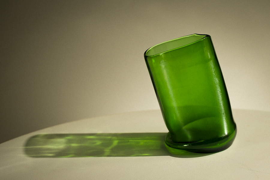 A translucent green glass tumbler or vase-like cup, shown standing on a white surface with a long green shadow cast against a warm beige background.