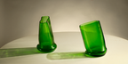 Two green translucent glass vases with a modern sculptural, tapered design, displayed on a white surface against a neutral gray background.