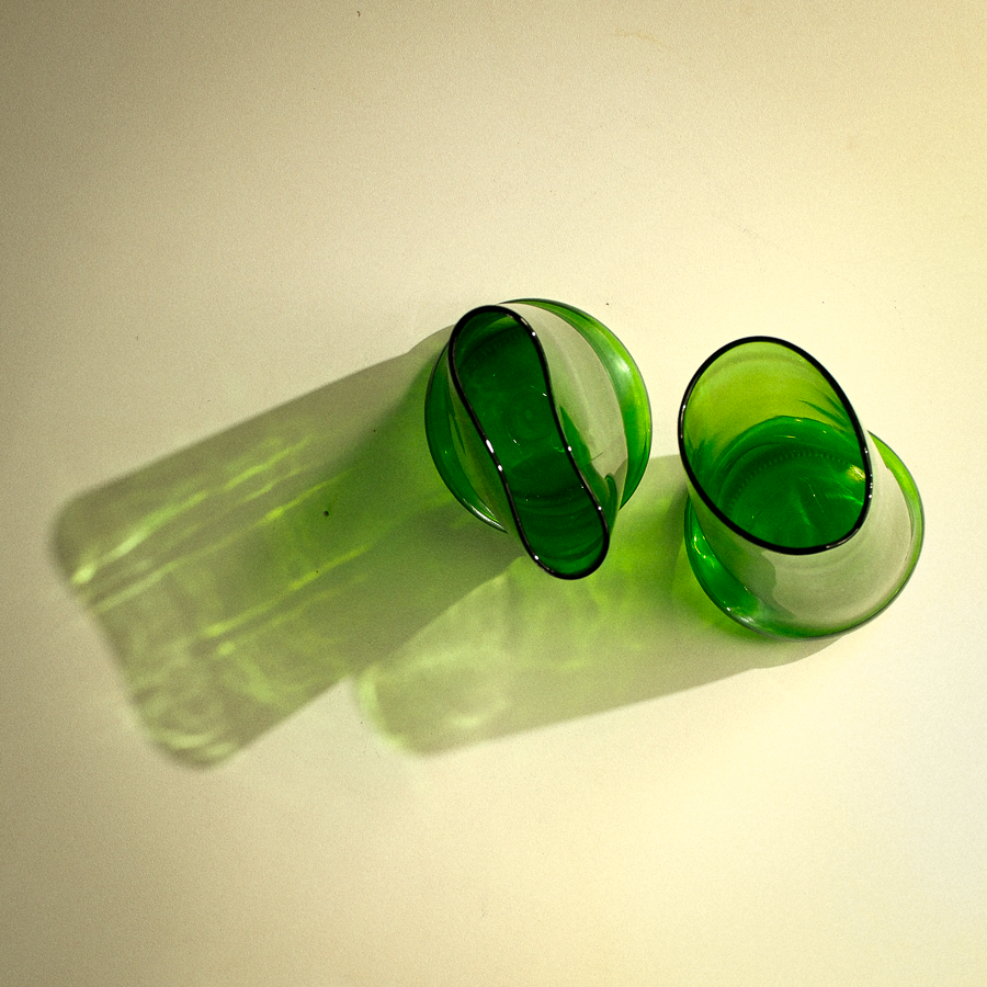Two green tinted glass shot glasses with black rims, shown on a warm off-white background with a strong shadow.