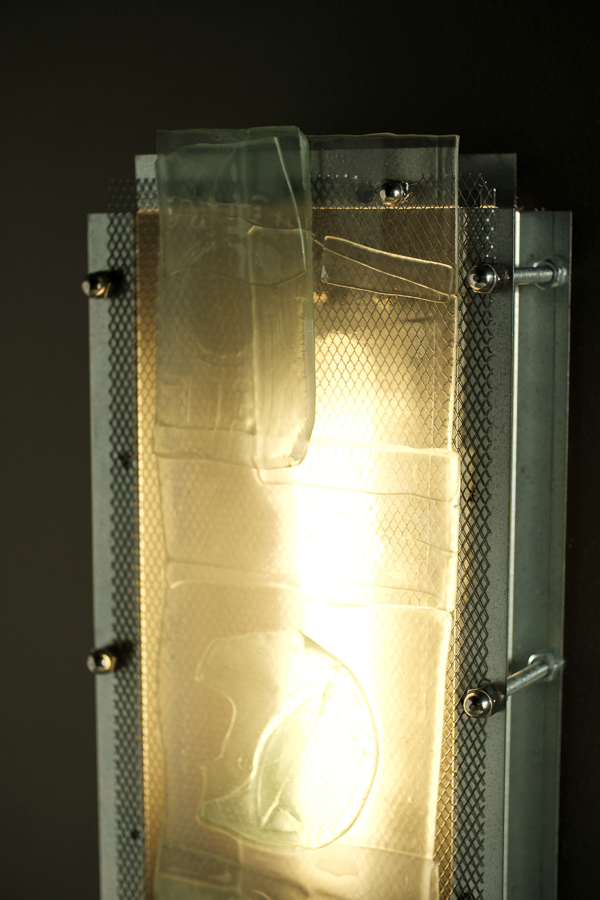 A rectangular translucent glass panel mounted on a metal frame with visible screws and mesh backing, glowing with warm light against a dark background.