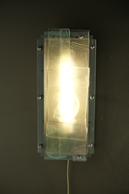 A rectangular wall-mounted light fixture with a glowing warm white bulb inside a clear textured glass panel, mounted on a dark wall.