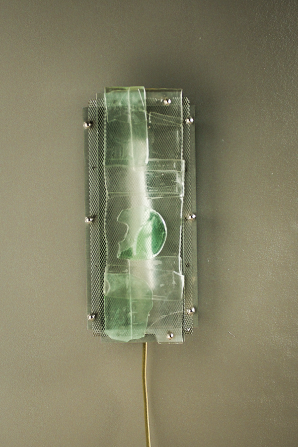 A rectangular wall-mounted light fixture with a translucent green glass shade, a white diffuser, and a thin yellow cord extending downward, shown against a plain taupe wall.