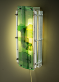A modern wall-mounted light fixture with a translucent green and yellow acrylic panel, a silver metal mesh screen, and visible mounting hardware, casting a warm yellow glow on the wall behind it.