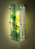 A modern wall-mounted light fixture with a translucent green and yellow acrylic panel, a silver metal mesh screen, and visible mounting hardware, casting a warm yellow glow on the wall behind it.