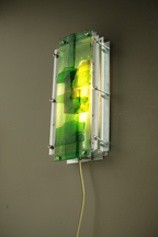 A modern wall-mounted light fixture with translucent green and yellow acrylic panels set in a silver metal frame, shown mounted on a dark gray wall.